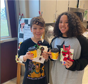  2 students holding their monster sewing projects and cocoa cups 